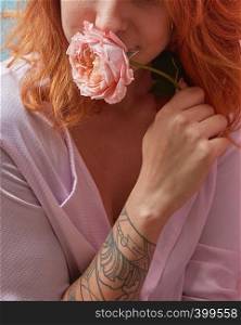 Fresh fragrant living coral colored rose in a girl's hands with tattoo. Smiling young pretty woman with ginger hair. Creeting card of Woman's or Mother's Day. Close-up.. Smiling happy red-haired girl in a pink shirt and delicate rose in her hand with tattoo copy space. Close-up. The gift for Mother's Day.