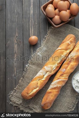 Fresh french baguette made from white flour next to chicken eggs and flour, ingredients for baking. Bread located on burlap, rustic dark wooden background. Top view. Vertical frame