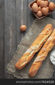 Fresh french baguette made from white flour next to chicken eggs and flour, ingredients for baking. Bread located on burlap, rustic dark wooden background. Top view. Vertical frame