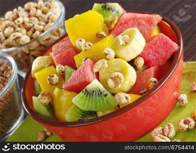 Fresh fruit salad made of banana, kiwi, watermelon and mango pieces in orange bowl with cereals (puffed wheat and puffed chocolate quinoa) (Selective Focus, Focus on the front of the bowl and the fruits in the front). Fruit Salad with Cereals