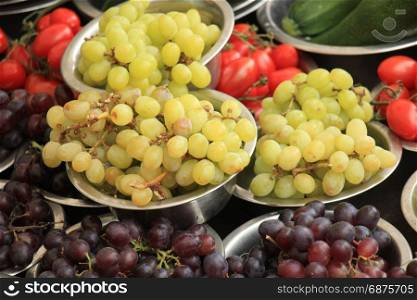 Fresh fruits and vegetables on a market stall: grapes, tomatoes, courgette and peppers displayed in small metal bowls