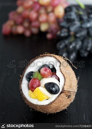 Fresh fruits are located in the half of coconut. Grape clusters in the background. Fruits are located on a black slate stone.. Fresh fruits in half a coconut on a black textured stone background, healthy nutrition, vegetarian diet.