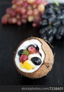 Fresh fruits are located in the half of coconut. Grape clusters in the background. Fruits are located on a black slate stone.. Fresh fruits in half a coconut on a black textured stone background, healthy nutrition, vegetarian diet.