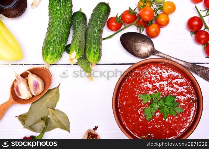 Fresh gazpacho soup in a brown ceramic dish with a spoon on a white wooden background, next to fresh vegetables and spices for cooking, top view