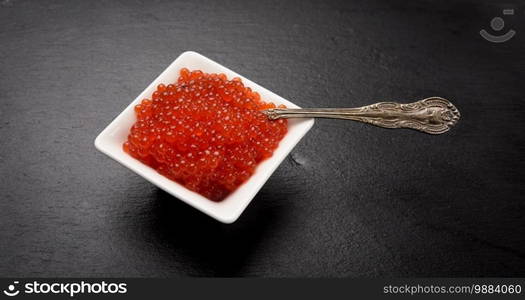 fresh grained red chum salmon caviar in a white ceramic bowl on a black slate stone background, close up