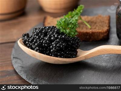 fresh grainy black paddlefish caviar in brown wooden spoon on a black background, close up