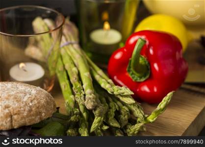 Fresh green asparagus spears with red bell pepper. Fresh green asparagus spears with red bell pepper lying on a wooden kitchen counter with lighted candles and fresh brown rolls