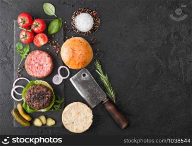 Fresh grilled and raw minced pepper beef burger on stone chopping board with buns onion and tomatoes on black background. Salty pickles and basil. Top view