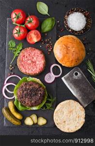 Fresh grilled and raw minced pepper beef burger on stone chopping board with buns onion and tomatoes on black background. Salty pickles and basil. Top view