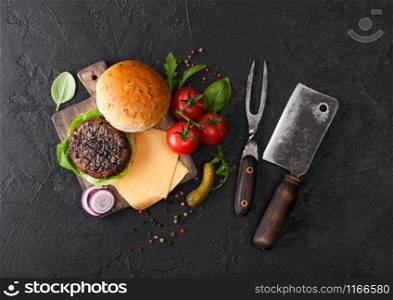 Fresh grilled and raw minced pepper beef burger on vintage chopping board with buns onion and tomatoes on black background. Salty pickles and basil with meat hatchet. Top view