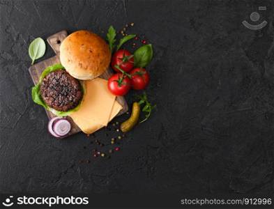 Fresh grilled and raw minced pepper beef burger on vintage chopping board with buns onion and tomatoes on black background. Salty pickles and basil. Top view