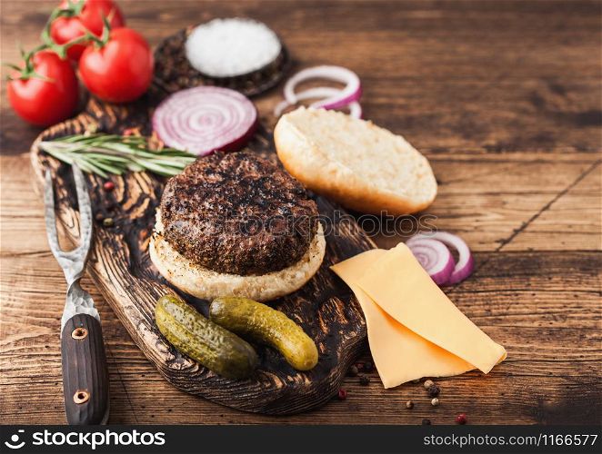 Fresh grilled minced pepper beef burger on vintage chopping board with buns onion and tomatoes on wood background. Salty pickles and fork