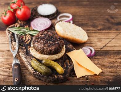 Fresh grilled minced pepper beef burger on vintage chopping board with buns onion and tomatoes on wood background. Salty pickles and fork