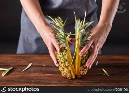 Fresh halves of an exotic pineapple hold the girl&rsquo;s hands on a wooden brown table with copy space. Vitamin fruit. Woman&rsquo;s hands hold pineapple halves with green leaves on a wooden brown table with copy space. Healthy product