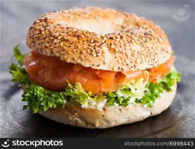 Fresh healthy bagel sandwich with salmon, ricotta and lettuce in black plate on dark kitchen table background.