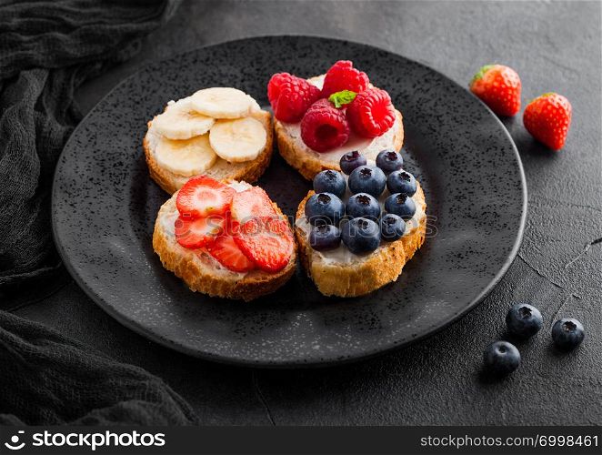 Fresh healthy mini sandwiches with cream cheese, fruits and berries in black with cloth. Strawberries, blueberries, bananas and raspberries on stone kitchen table background.