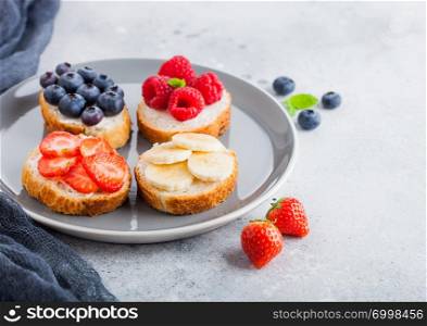 Fresh healthy mini sandwiches with cream cheese, fruits and berries in grey plate with cloth. Strawberries, blueberries, bananas and raspberries on stone kitchen table background.