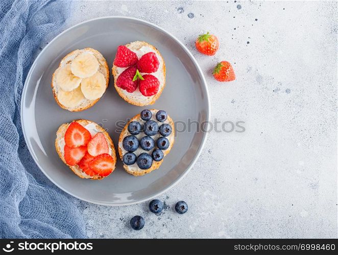 Fresh healthy mini sandwiches with cream cheese, fruits and berries in grey plate with cloth. Strawberries, blueberries, bananas and raspberries on stone kitchen table background.