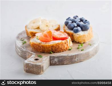 Fresh healthy mini sandwiches with cream cheese, fruits and berries on vintage wooden board. Strawberries, blueberries, bananas and raspberries on stone kitchen table background.