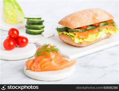 Fresh healthy salmon sandwich with lettuce and cucumber and tomatoes on marble board on white marble background.Diet healthy food