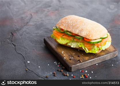 Fresh healthy salmon sandwich with lettuce and cucumber on vintage chopping board on black stone background. Breakfast snack