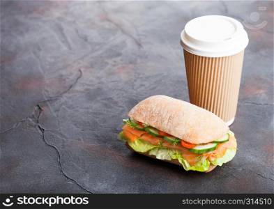 Fresh healthy salmon sandwich with lettuce and cucumber with paper cup of coffee on black stone background. Breakfast snack.