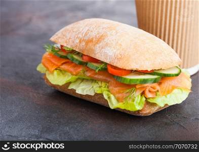 Fresh healthy salmon sandwich with lettuce and cucumber with paper cup of coffee on black stone background.