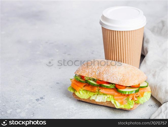 Fresh healthy salmon sandwich with lettuce and cucumber with paper cup of coffee on white stone background. Breakfast snack.