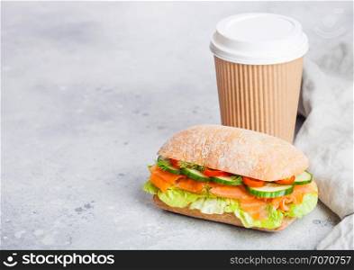 Fresh healthy salmon sandwich with lettuce and cucumber with paper cup of coffee on white stone background. Breakfast snack.