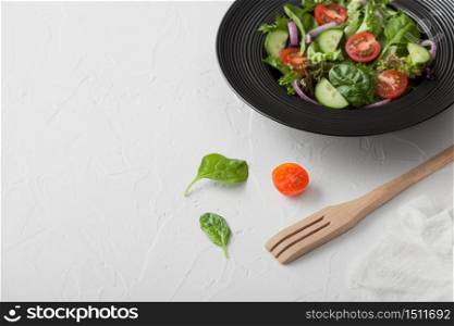 Fresh healthy vegetarian vegetables salad with lettuce and tomatoes, red onion and spinach in black bowl on white background with spatula fork and white kitchen cloth. Top view