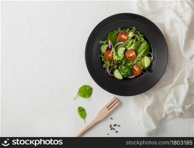 Fresh healthy vegetarian vegetables salad with lettuce and tomatoes, red onion and spinach in black bowl on white background with spatula fork and white kitchen cloth. Top view