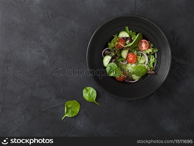 Fresh healthy vegetarian vegetables salad with tomatoes and cucumber, red onion and spinach in black bowl on dark table background. Top view.