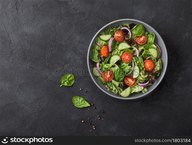 Fresh healthy vegetarian vegetables salad with tomatoes and cucumber, red onion and spinach in grey bowl plate on dark background. Top view.