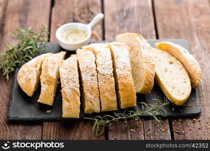 fresh homemade ciabatta bread sliced on stone slate board, olive oil and rosemary on wooden background