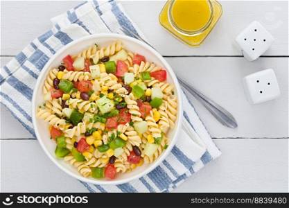 Fresh homemade colorful vegan fusilli pasta salad with beans, corn, tomato, cucumber and green bell pepper served in bowl, vinaigrette dressing on the side, photographed overhead on white wood (Selective Focus, Focus on the salad). Vegan Fusilli Pasta Salad
