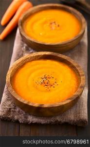 Fresh homemade cream of carrot soup with freshly ground black pepper on top served in wooden bowls, photographed on dark wood with natural light (Selective Focus, Focus in the middle of the first soup)