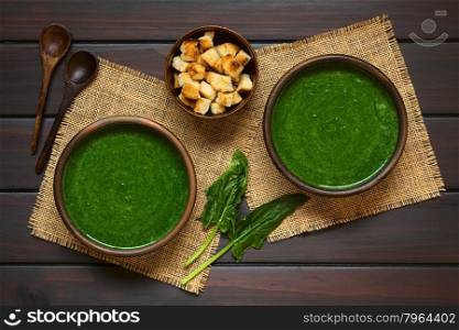 Fresh homemade cream of spinach soup in rustic bowls, homemade croutons, wooden spoons and fresh spinach leaves on the side, photographed overhead on dark wood with natural light
