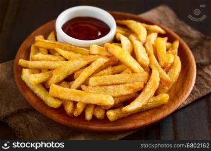 Fresh homemade crispy French fries with a small bowl of ketchup on wooden plate, photographed with natural light (Selective Focus, Focus one third into the fries)