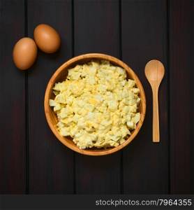 Fresh homemade egg salad prepared with mayonnaise and mustard in wooden bowl, with raw eggs and wooden spoon on the side, photographed overhead on dark wood with natural light