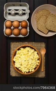 Fresh homemade egg salad prepared with mayonnaise and mustard in wooden bowl, with egg carton and slices of wholegrain bread above, photographed overhead on dark wood with natural light