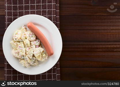 Fresh homemade German Kartoffelsalat potato salad made of potato, onion, pickle and mayonnaise, served with sausage on plate, photographed with copy space overhead on dark wood. Potato Salad with Sausage