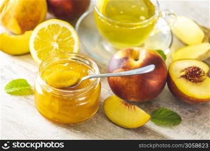 Fresh homemade peach jam in glass jar on a light gray wooden background. Several fresh berries, lemon and mint are near it.