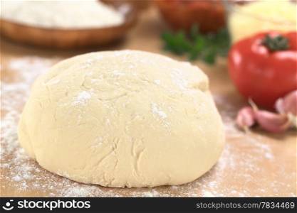 Fresh homemade pizza dough on floured wooden board with pizza ingredients (flour, tomato, grated cheese and herbs) in the back (Selective Focus, Focus one third into the pizza dough) . Pizza Dough