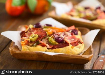Fresh homemade pizza piece with tomato sauce, ham, salami, olives, bell pepper and cheese on top, served on sandwich paper on wooden plate (Selective Focus, Focus on the front of the olive slices)