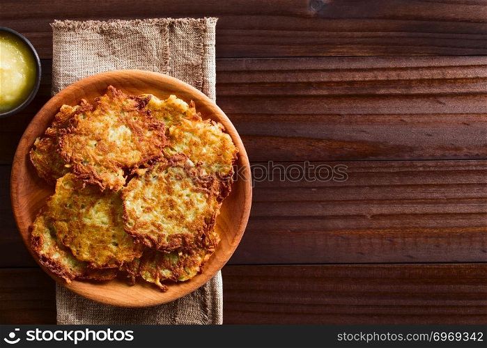 Fresh homemade potato fritters or pancakes on wooden plate with apple sauce on the side, a traditional German snack or dish called Kartoffelpuffer or Reibekuchen, photographed overhead on dark wood with copy space on the side. Fresh Homemade Potato Fritters or Pancakes