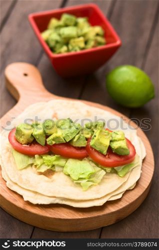 Fresh homemade tortilla with lettuce, tomato and avocado on wooden board with avocado salad and lime in the back (Selective Focus, Focus on the front of the avocado pieces on top)