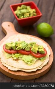 Fresh homemade tortilla with lettuce, tomato and avocado on wooden board with avocado salad and lime in the back (Selective Focus, Focus on the front of the avocado pieces on top)