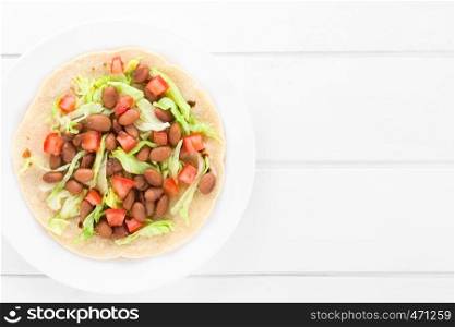 Fresh homemade vegan tacos made of flour tortilla with lettuce, cooked beans and tomatoes served on plate, photographed overhead on white wood with copy space on the right side. Flour Tortilla with Beans, Lettuce and Tomato