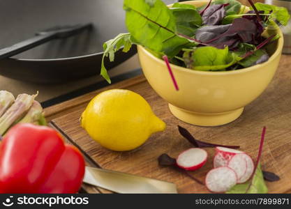 Fresh ingredients for making a salad on a chopping board in the kitchen with sliced radish, rocket, baby spinach and herbs in a bowl, red bell pepper and a ripe yellow lemon