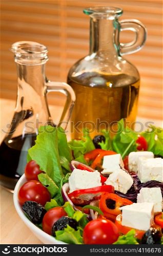 Fresh lettuce, tomato, red pepper, red onion, black olive and feta cheese salad with bottles of olive oil and balsamic vinegar dressing in the background.